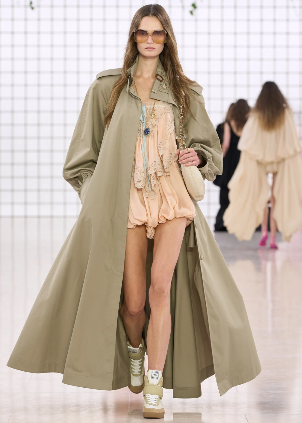 Desfile de moda apresenta look com trench coat bege longo, vestido curto em tons pastel com detalhes em renda e tênis esportivo. A modelo usa óculos de sol oversized e carrega bolsa de ombro. A combinação destaca a mistura de elementos clássicos e casuais, tendência atual.