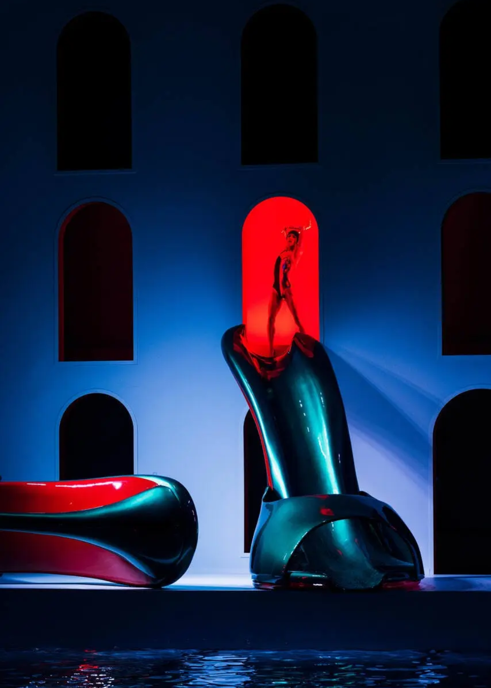 Cena de desfile de moda futurista com iluminação dramática em azul e vermelho. O modelo traja um body estilizado brilhante seguindo tendências modernas e ousadas, posicionado em uma estrutura de design contemporâneo iluminada, destacando a combinação de moda e arte.