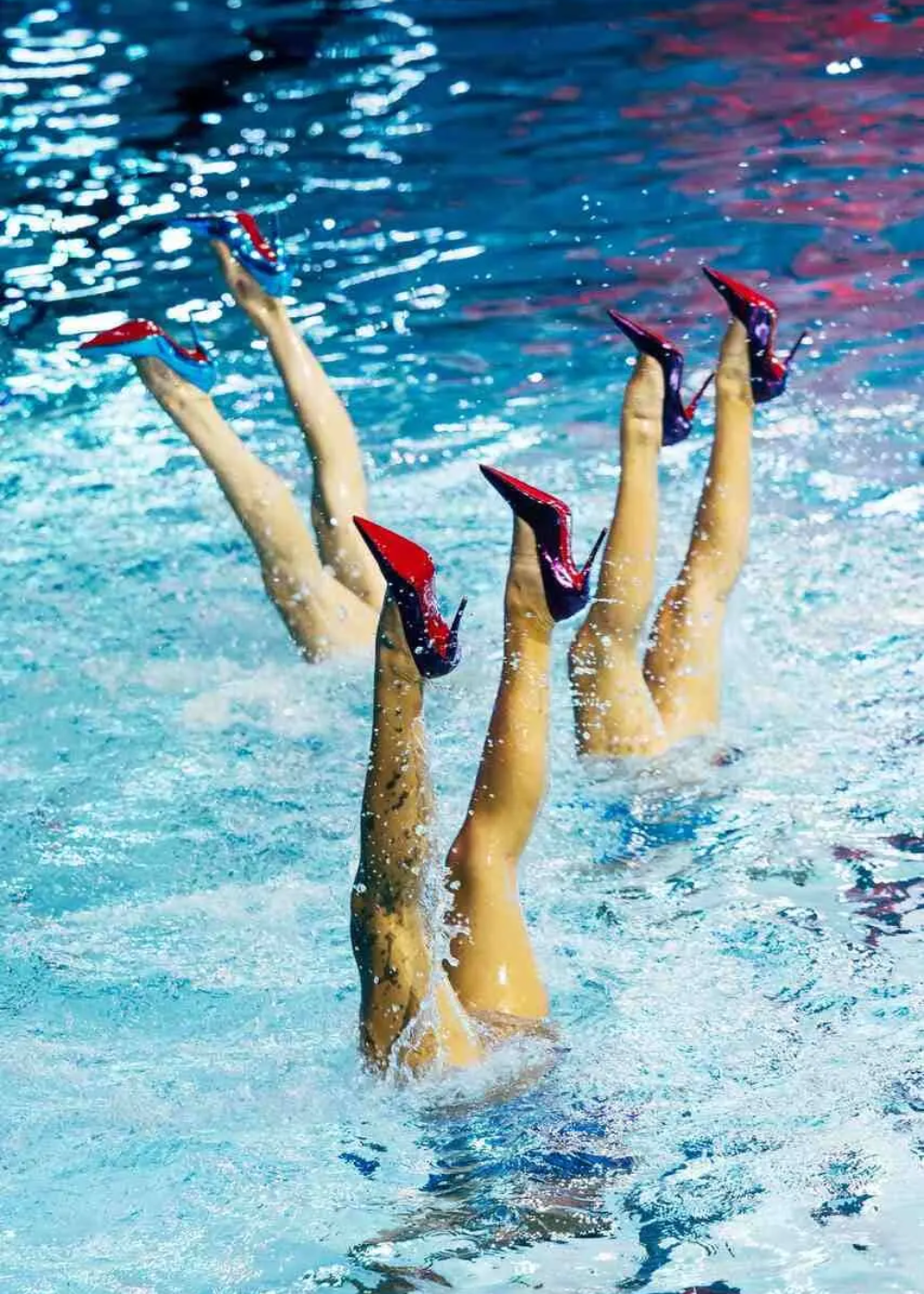 Imagem de pernas submersas em coreografia de nado sincronizado, todas usando sapatos de salto alto vermelhos de verniz, tendência ousada e inusitada na moda aquática. Destaque para o contraste entre a elegância dos calçados e a dinâmica do movimento na água.