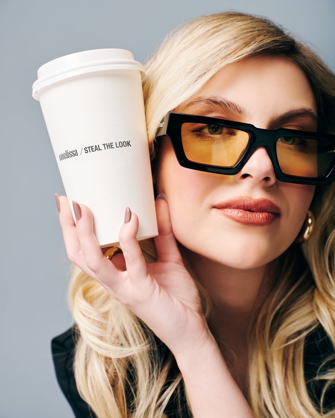 Mulher com óculos de sol retangulares modernos, cabelo loiro solto e maquiagem suave. Segura um copo de café com a marca 