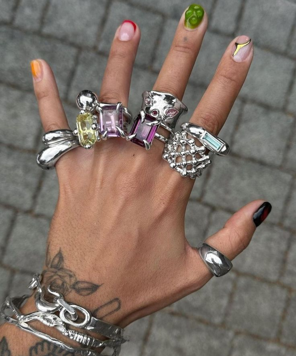 Mão tatuada exibindo várias tendências de moda: anéis prateados grandes e chamativos com pedras coloridas, pulseiras grossas prateadas, e unhas decoradas com nail art variada e cores vibrantes. Estilo moderno e arrojado.