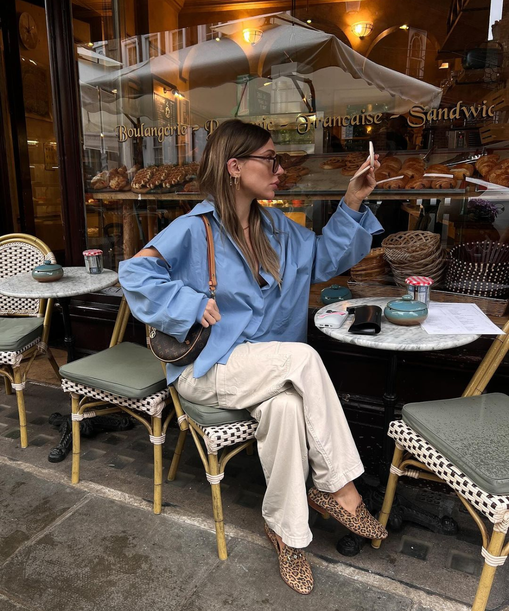 Mulher com camisa azul oversized, calça bege e sapato de oncinha, sentada em uma padaria francesa. Look casual e elegante, combinando tendências de conforto e estilo urbano. Bolsa pequena e óculos de sol completam o visual.