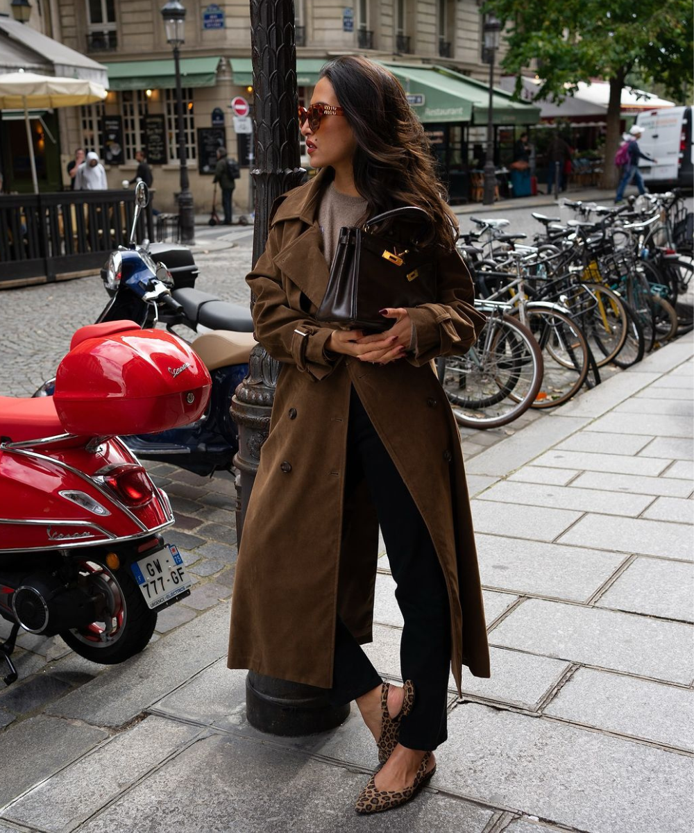 Mulher vestindo trench coat marrom, óculos de sol elegantes e sapato de oncinha em rua urbana. O look combina elegância clássica com toques modernos, perfeito para o outono. Tendência: sobreposições e acessórios refinados.