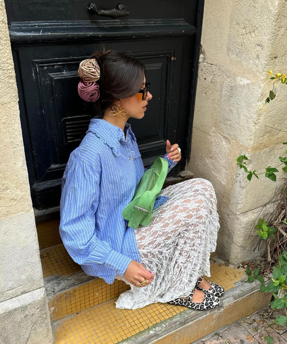 Pessoa com look casual eclético: camisa listrada azul oversized, saia de renda branca, bolsa verde vibrante e sapatos estampados. Acessórios chamativos incluem óculos de sol e brincos grandes. Cabelo preso com flores coloridas.