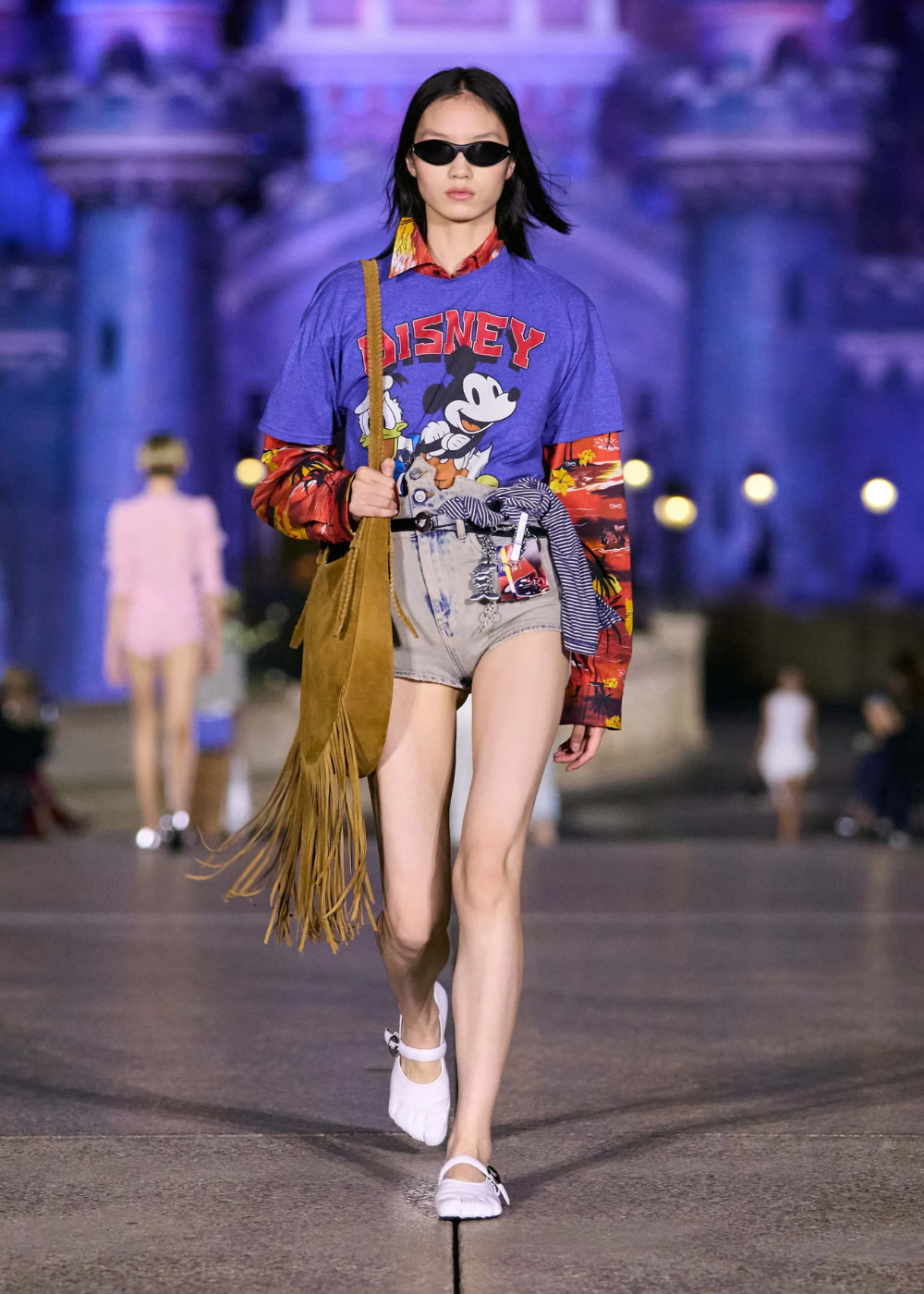 Modelo usa camiseta oversized temática da Disney sobre camisa de manga longa estampada. Shorts jeans de cintura alta com detalhes desbotados, bolsa de franjas e óculos de sol retrô complementam o look. Tendências: misturas de estampas e elementos retrô.