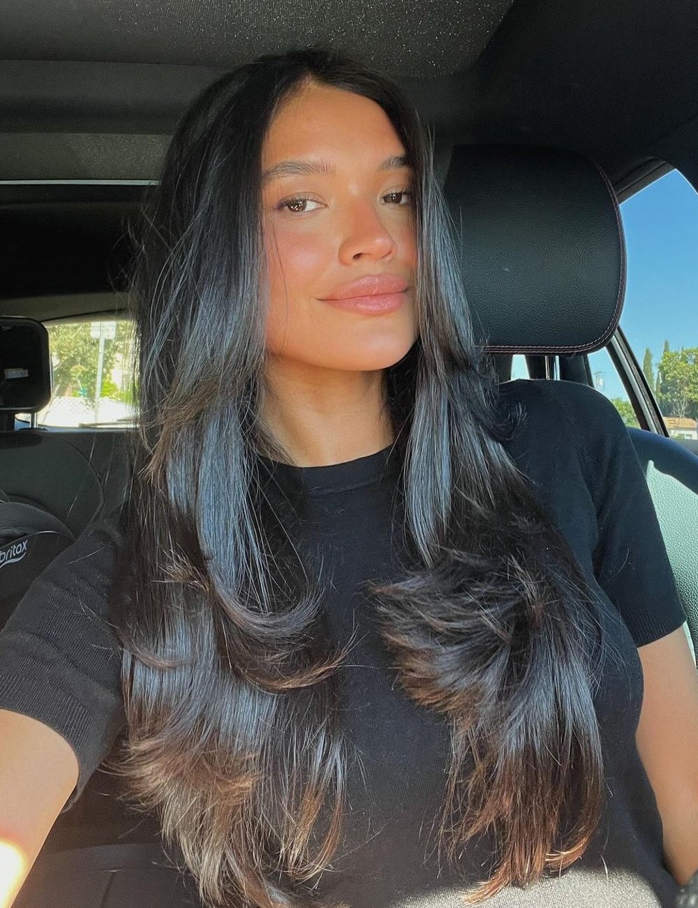 Mulher com cor de cabelo escuro, sentada dentro de um carro, usando camiseta preta.