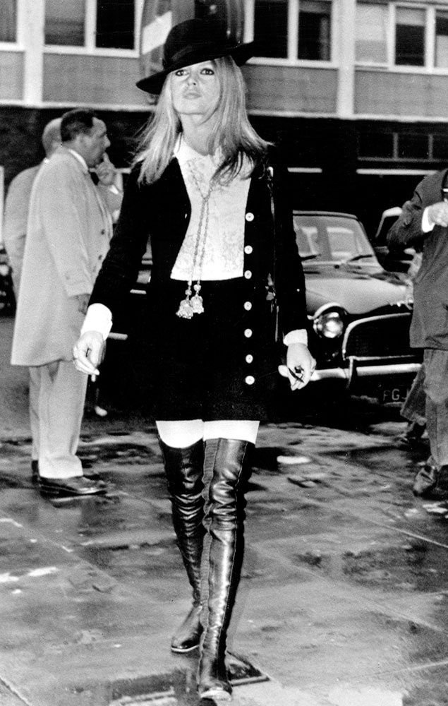 Mulher com chapéu e botas de couro, vestindo moda inspirada nos looks dos anos 70, caminha na rua.