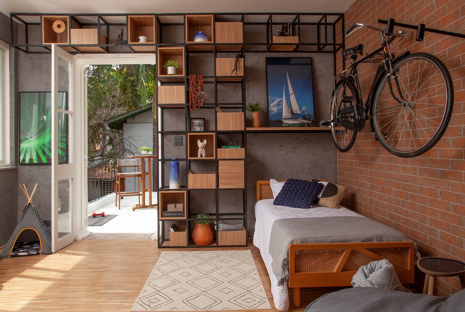 Quarto com decoração cinza e parede de tijolos. Estante de madeira moderna, bicicleta pendurada e cama com colcha cinza.