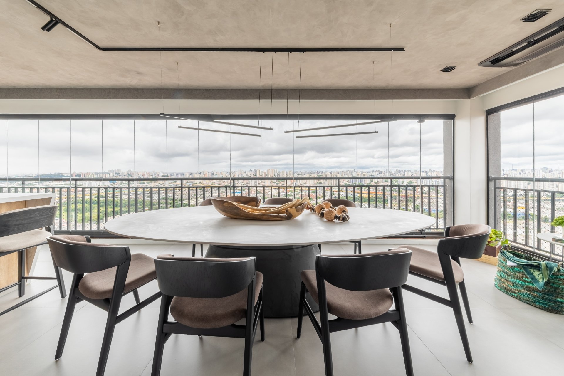 Sala de jantar elegante com mesa redonda cinza, cadeiras pretas e decoração natural, vista para a cidade ao fundo. Decoração cinza.