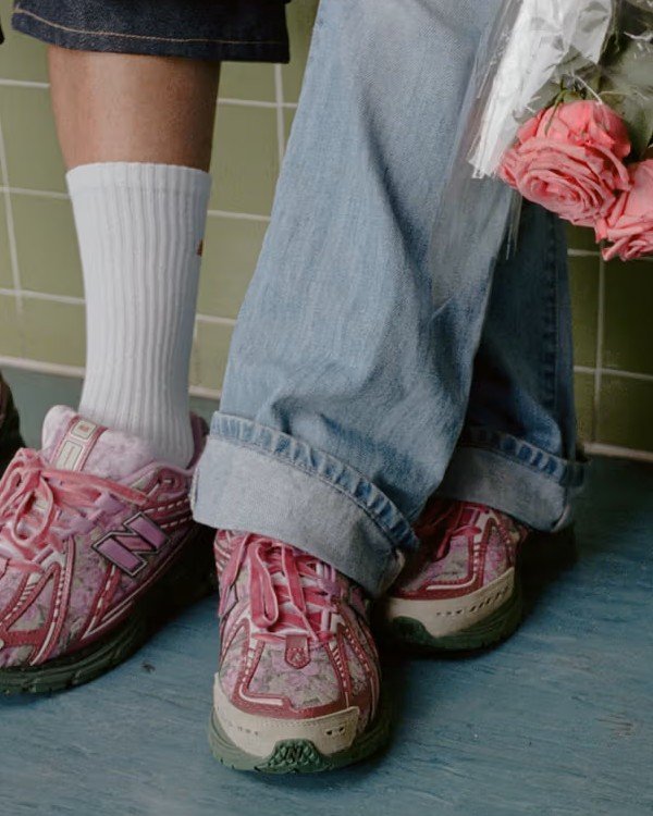 Tênis rosa com detalhes verdes sendo usados por pessoas com calças jeans e meias brancas. Uma delas segura rosas.