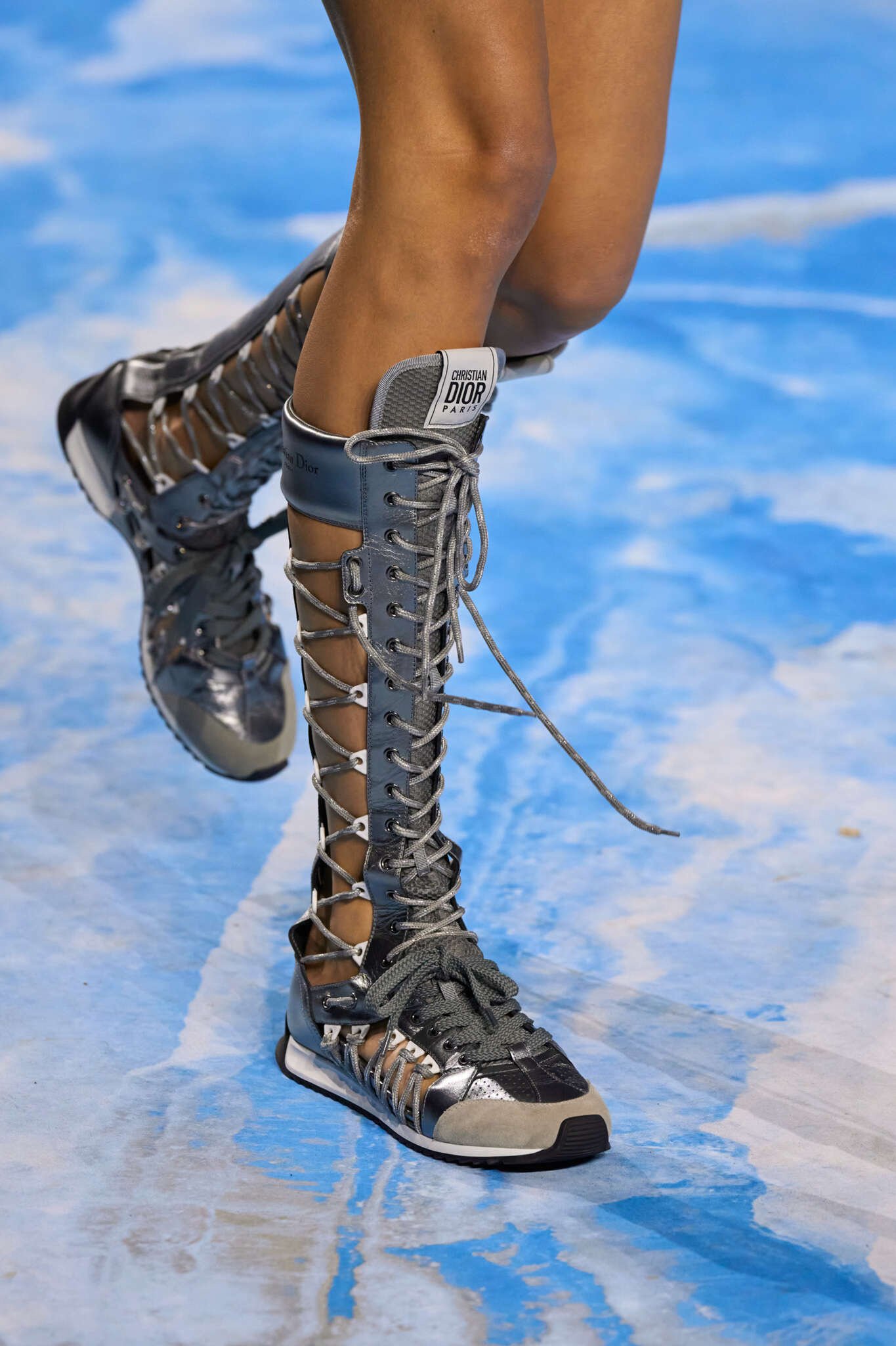 Botas prateadas de cano alto com cadarços, estilo boxing boots, em um desfile em passarela azul.