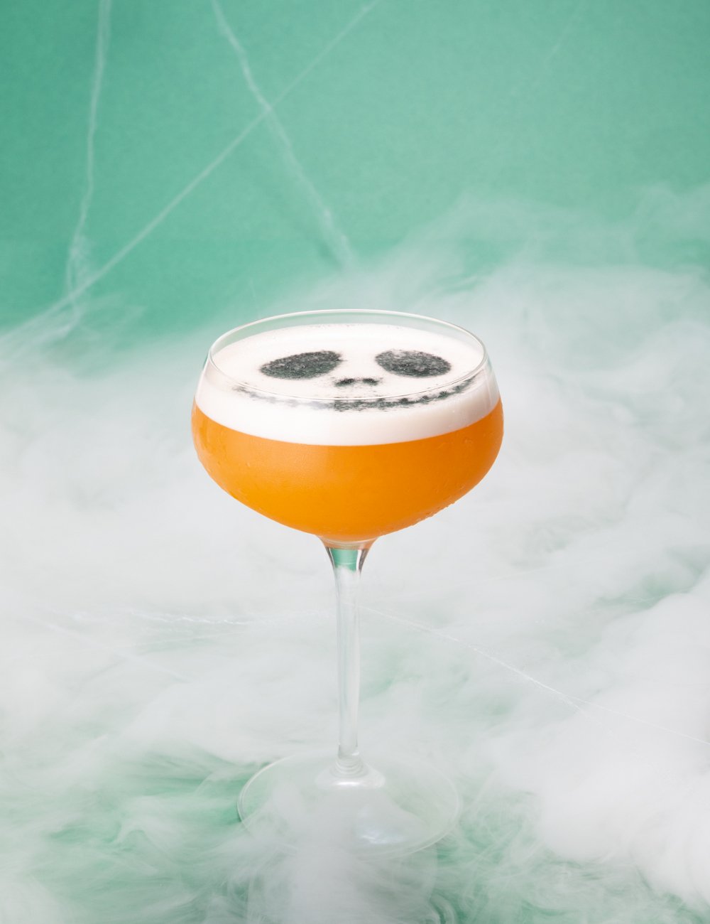 Coquetel laranja com espuma branca e desenho de caveira, cercado de fumaça.  Drinks de Halloween.