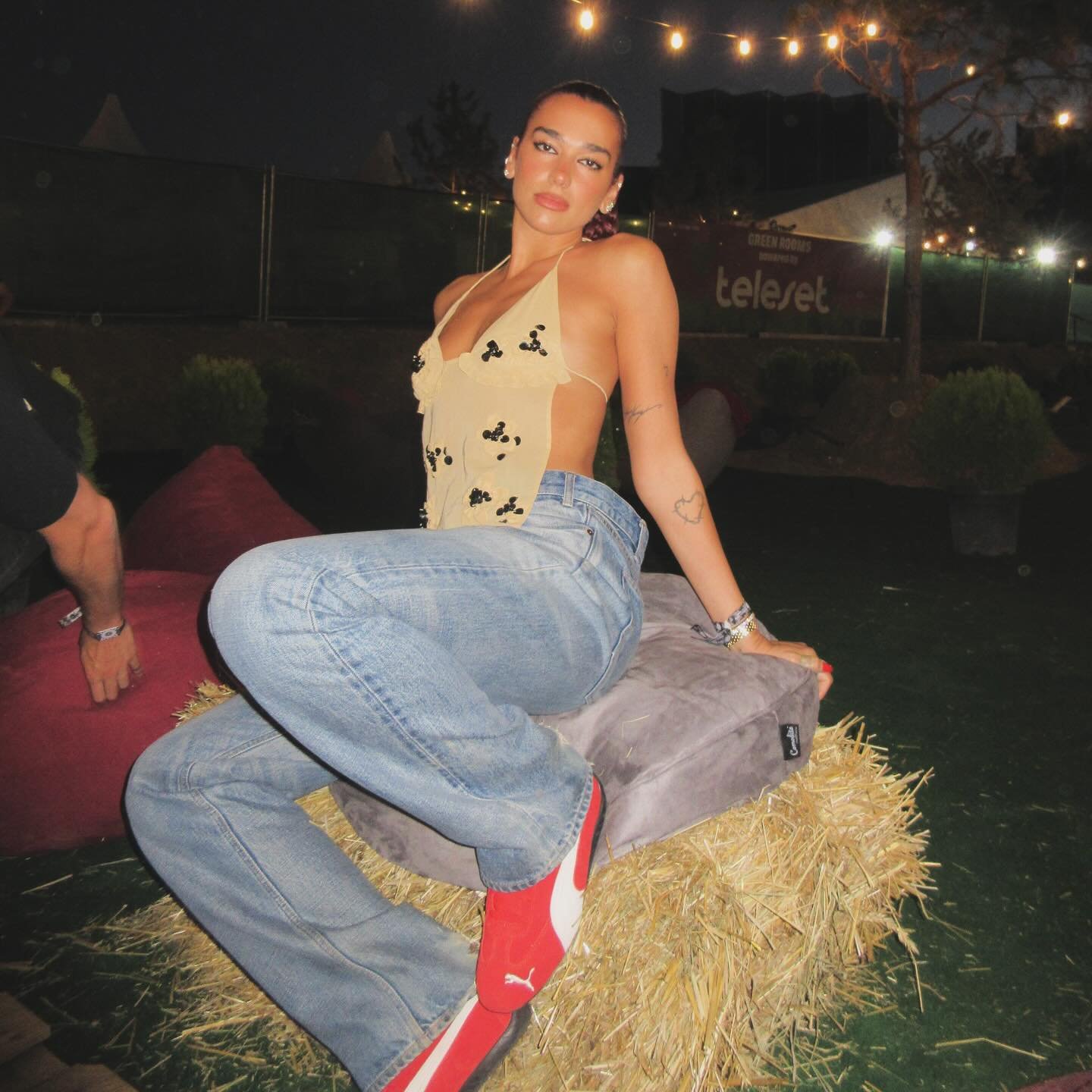 Pessoa sentada em feno ao ar livre à noite, usando top bege, jeans e tênis vermelho moderno.