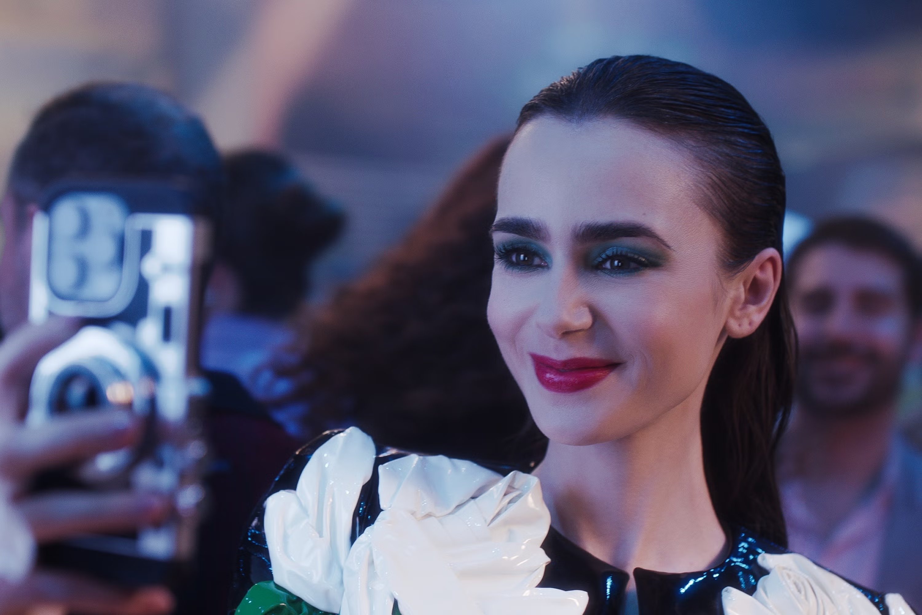 Jovem sorrindo tira selfie em evento, veste roupa futurista de vinil com detalhes florais brancos. Maquiagem marcante, olhos esfumados escuros e batom vermelho. Look ousado, refletindo tendências modernas e arrojadas. Lily Collins