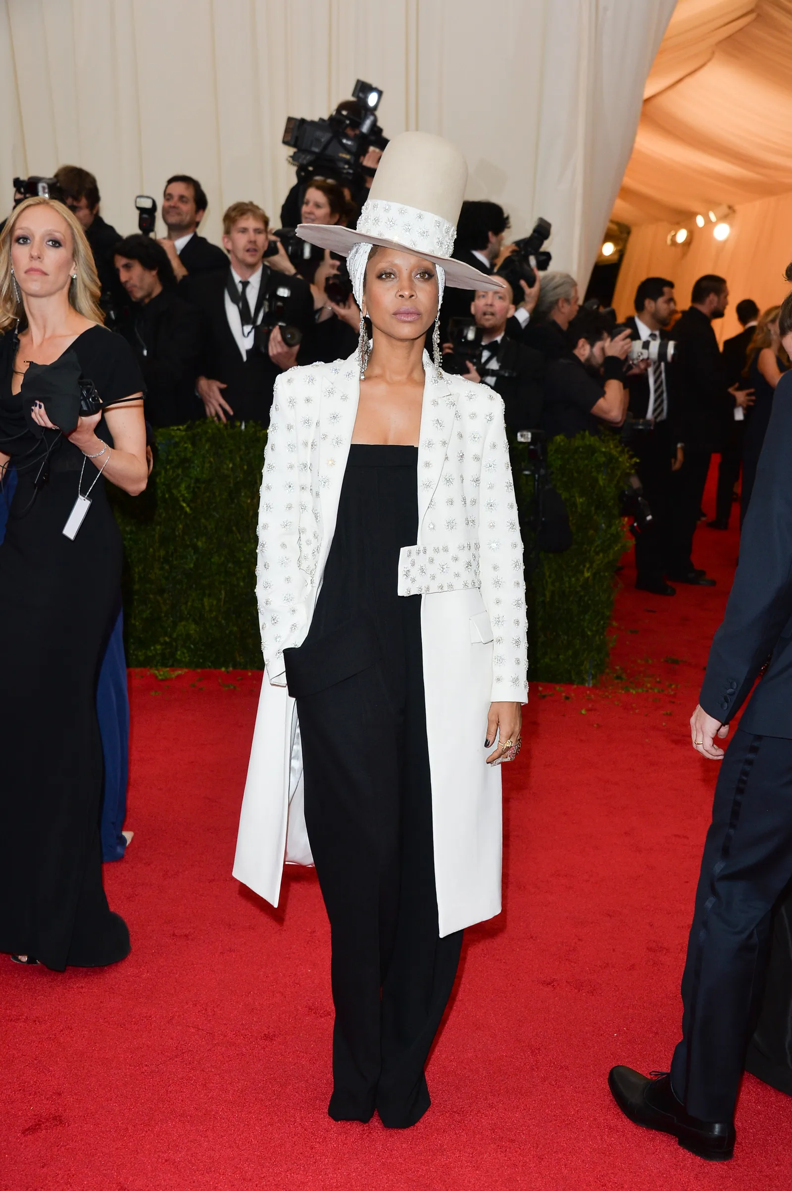 Na imagem, Erykah Badu usa um chapéu de aba larga estilizado e um casaco longo branco com detalhes bordados, sobre um conjunto preto. O look exala sofisticação e ousadia, destacando tendências de alta-costura e acessórios chamativos no tapete vermelho.