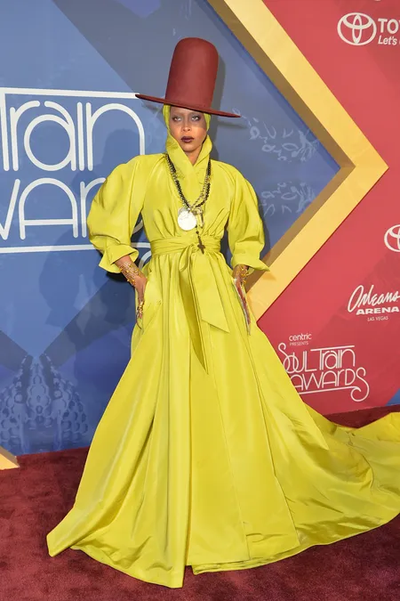 Figura feminina com vestido longo amarelo vibrante, de mangas bufantes, e cinto que acentua a cintura. Completa o visual com chapéu marrom de aba larga e colares. Look fashionista marcante e ousado, destacando-se em evento de premiação.