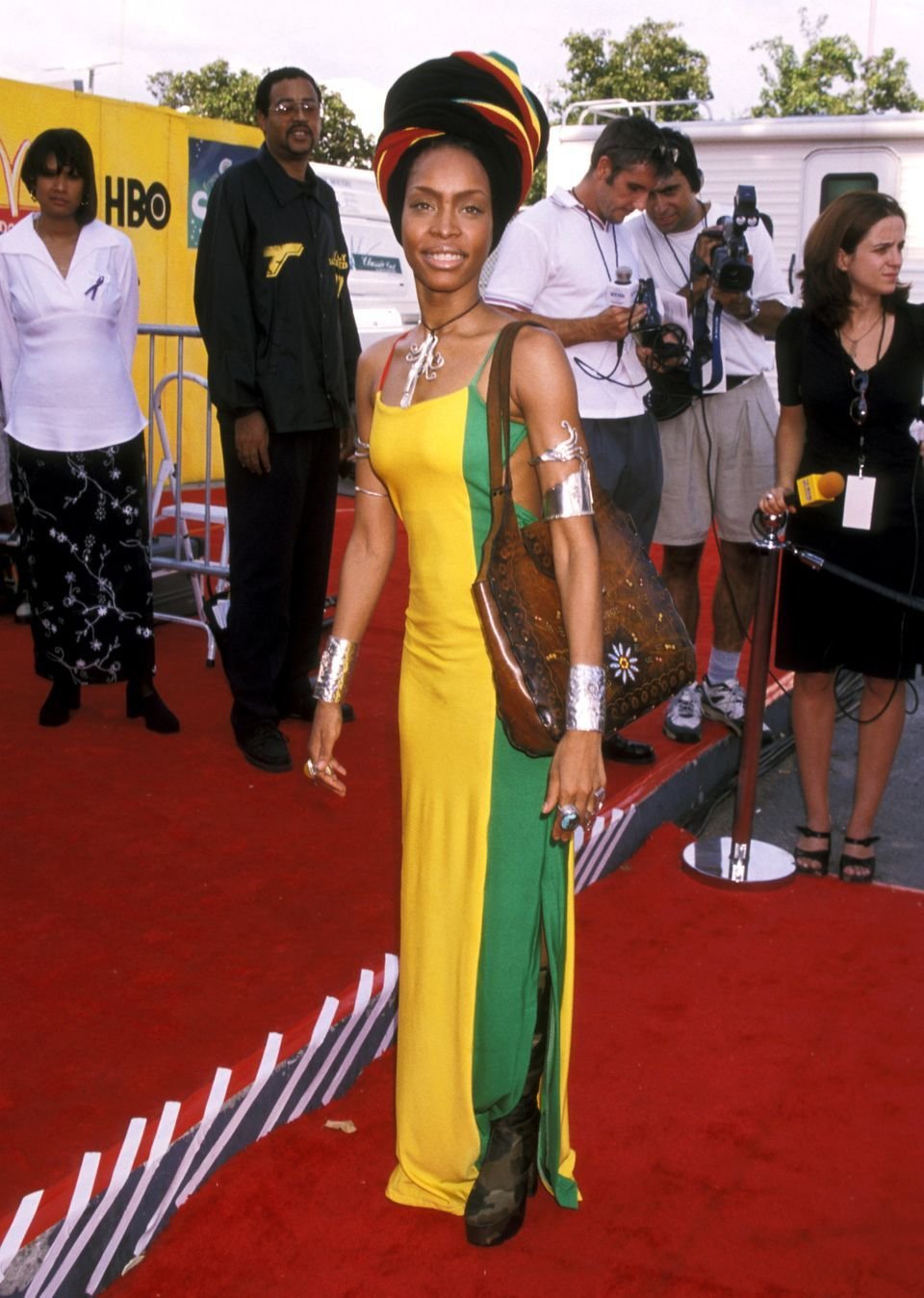 Imagem de um evento de tapete vermelho com uma mulher usando um vestido longo verde e amarelo, acessórios metálicos, botas e um turbante colorido. O estilo demonstra influências afro, com uma bolsa de couro artesanal completando o visual.