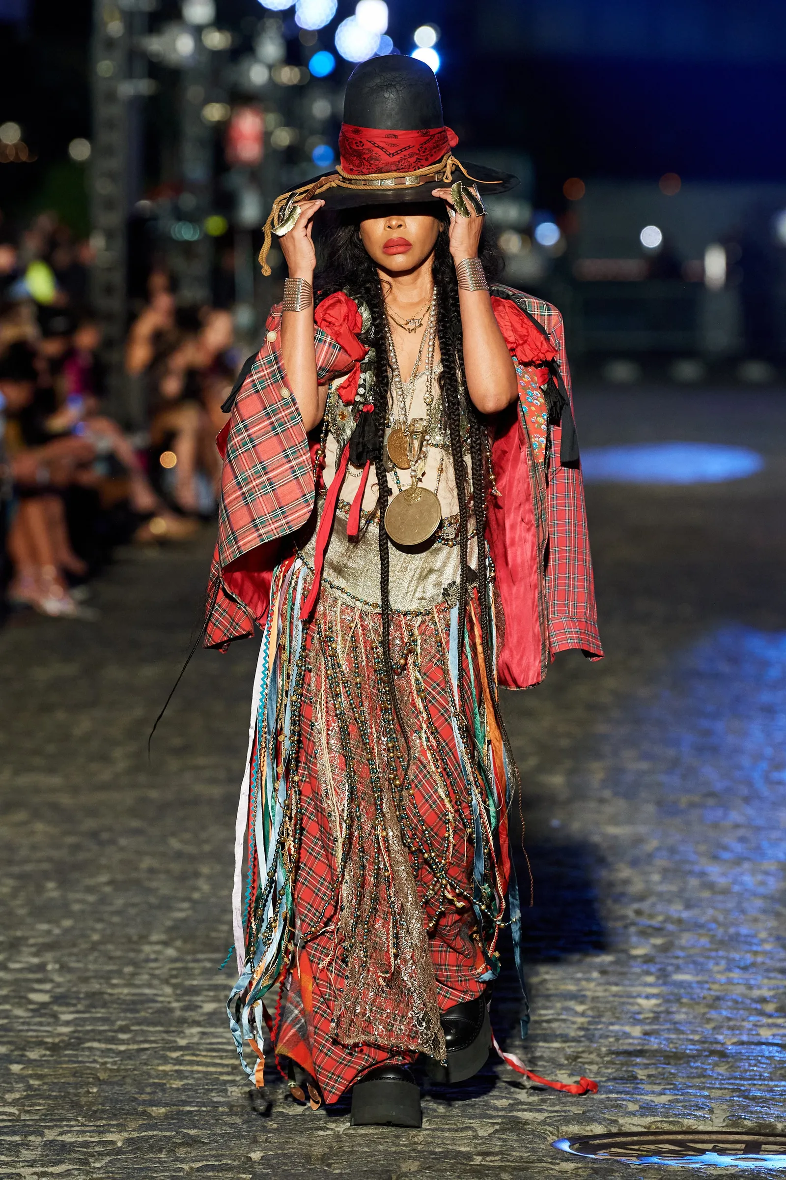 Desfile de moda com inspiração boho: modelo veste chapéu preto alto com lenço vermelho, casaco xadrez, blusa bege e saia longa com fitas coloridas. A variedade de texturas e acessórios reforça um visual eclético e artesanal.