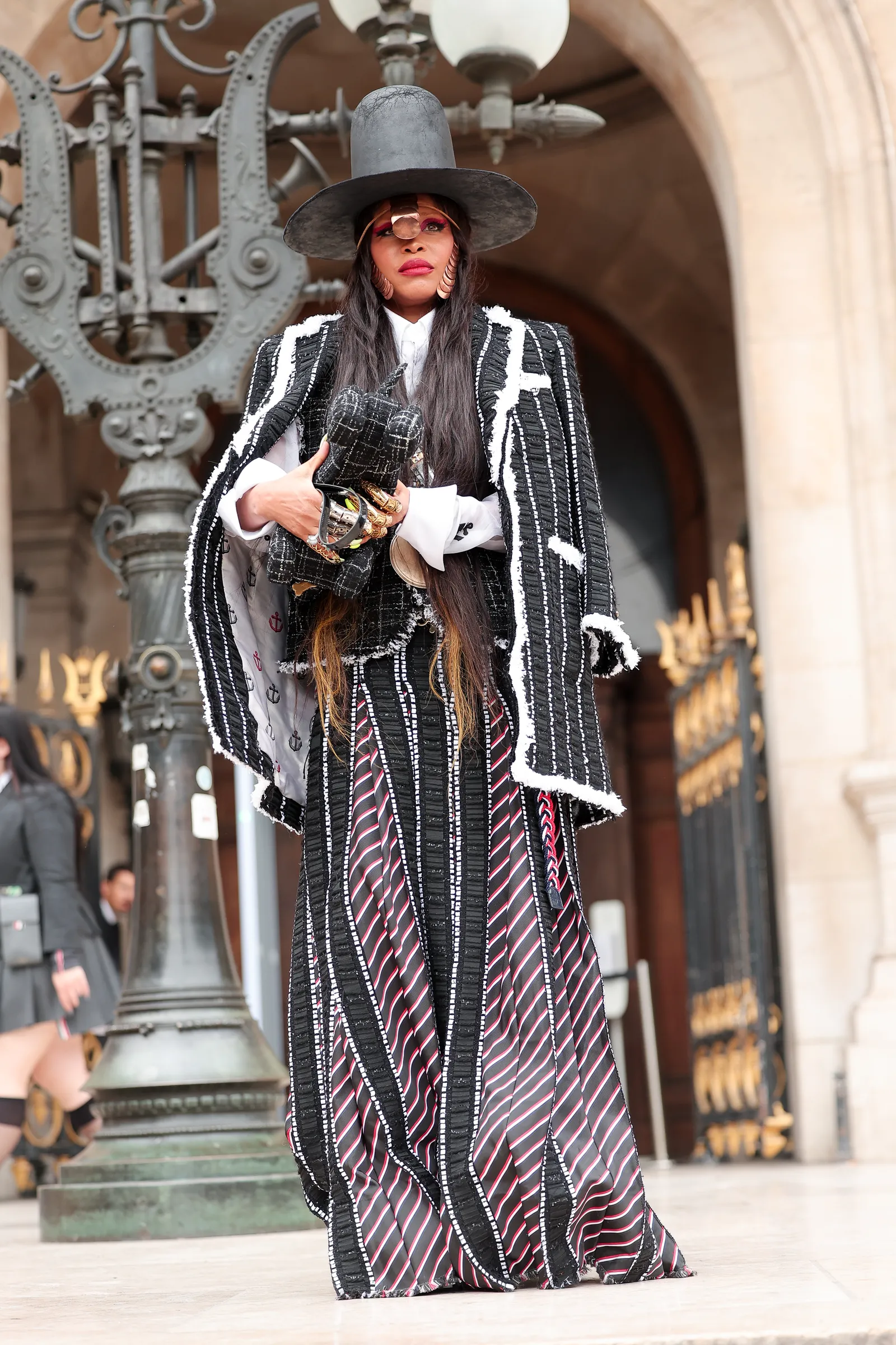 Pessoa vestida com um casaco preto xadrez estilo oversized, saia longa com listras pretas, vermelhas e brancas, e chapéu preto de aba larga. O visual mistura tendências contemporâneas e clássicas, com destaque para acessórios dourados e maquiagem marcante.