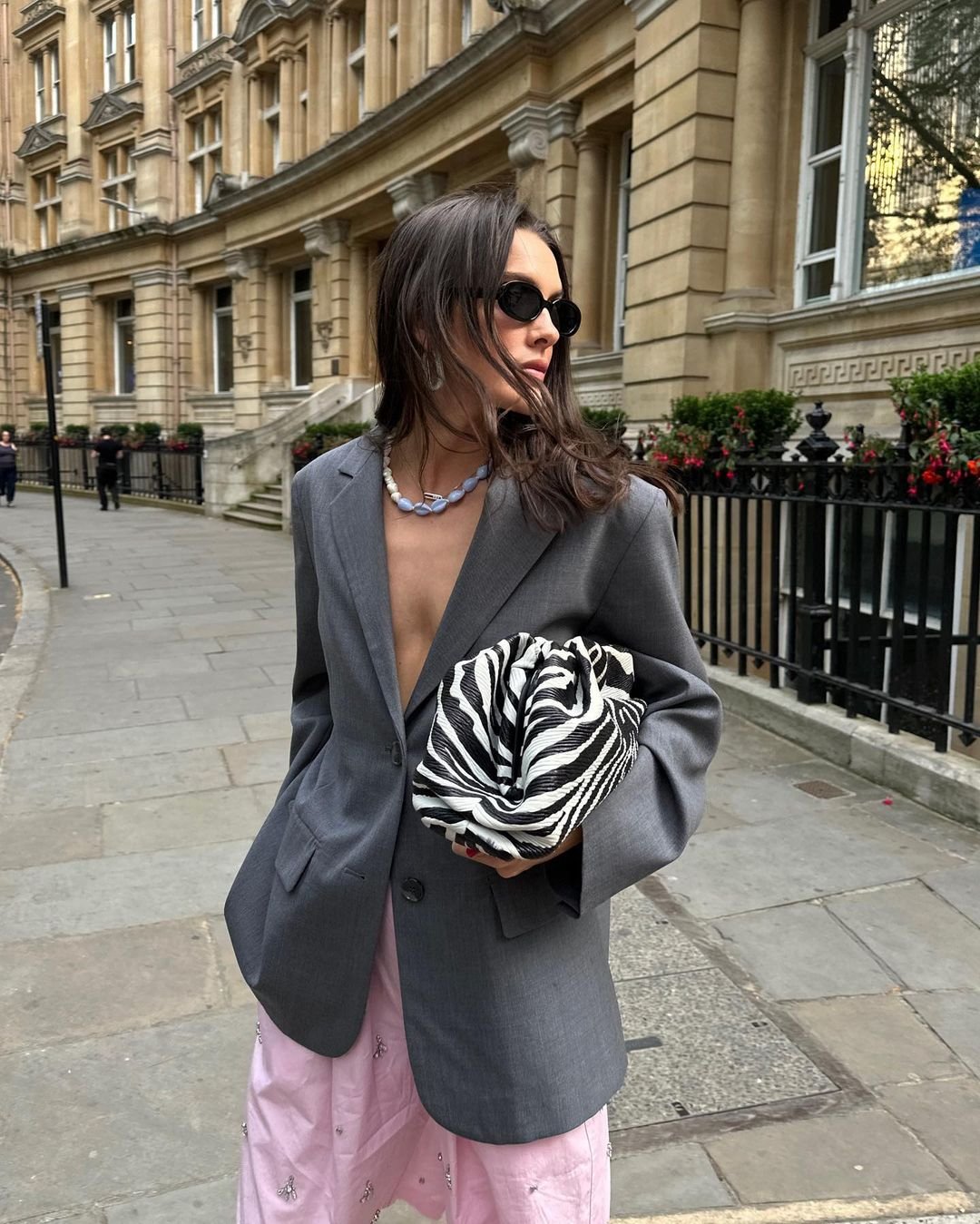 Modelo com blazer cinza oversized, calça rosa e bolsa com estampa de zebra, posa em rua com prédios clássicos ao fundo.