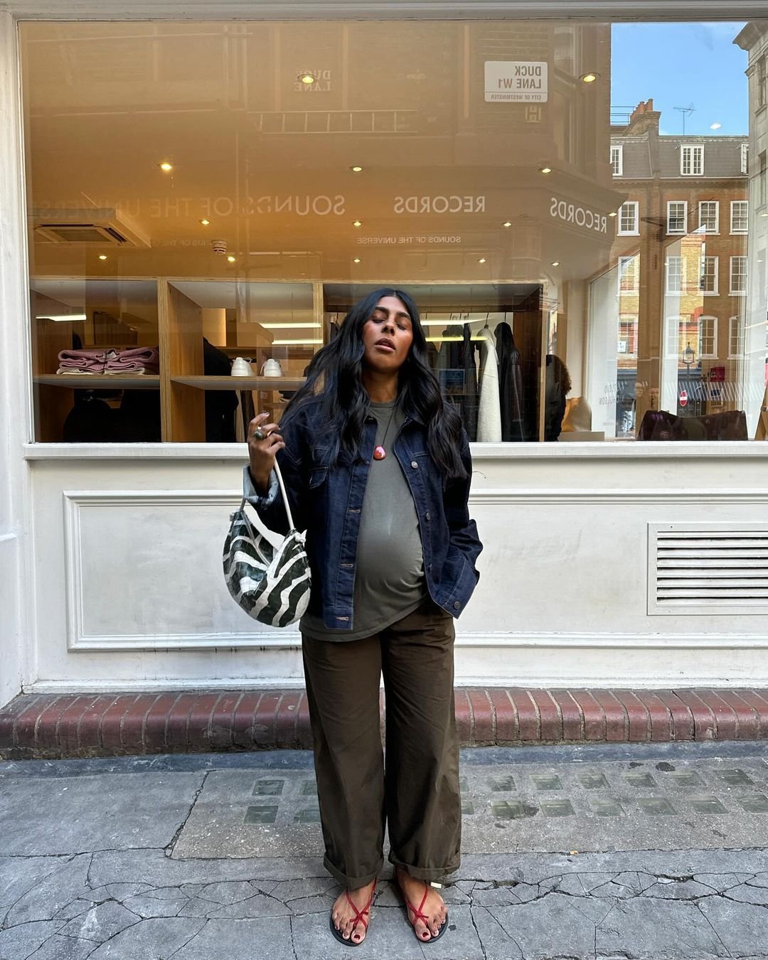 Mulher grávida usando jaqueta jeans, calça larga marrom e sandálias vermelhas, segura uma bolsa com estampa de zebra em frente a uma vitrine.