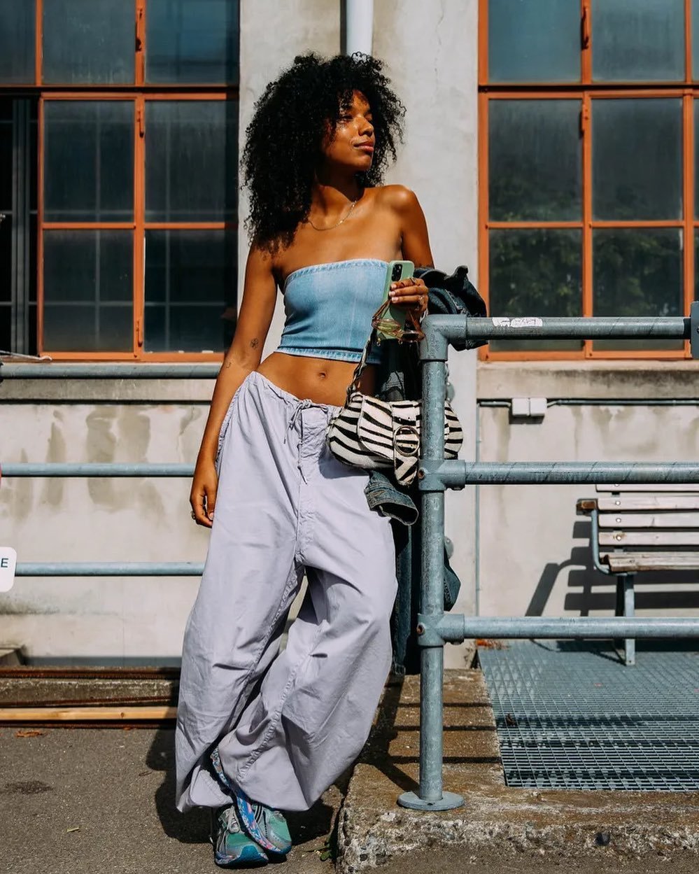 Mulher com top jeans, calça larga cinza e tênis, segura bolsa com estampa de zebra enquanto posa em frente a prédio com janelas laranjas.