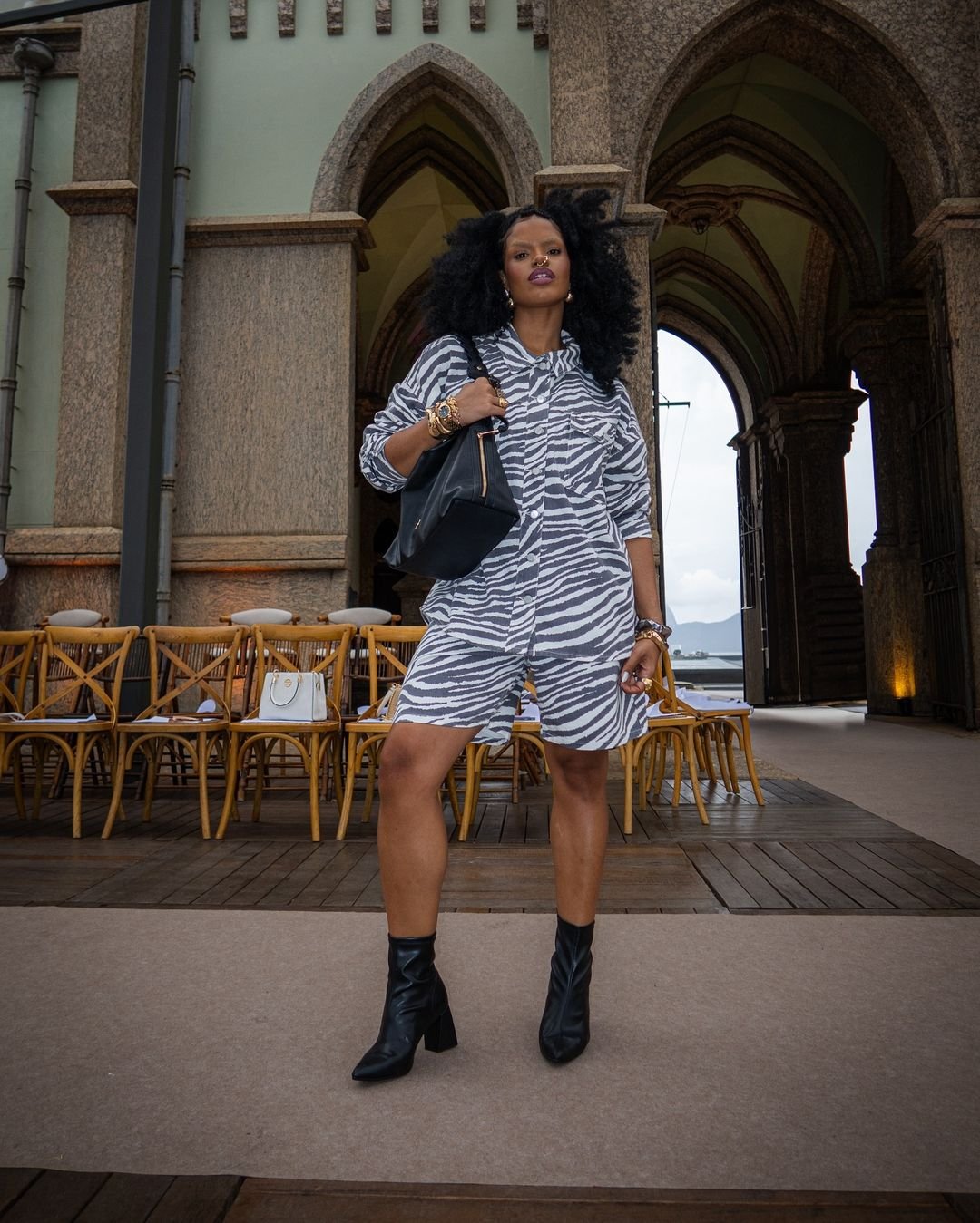 Mulher com vestido de estampa de zebra, botas pretas e bolsa de couro, posa em local histórico com arcos ao fundo.