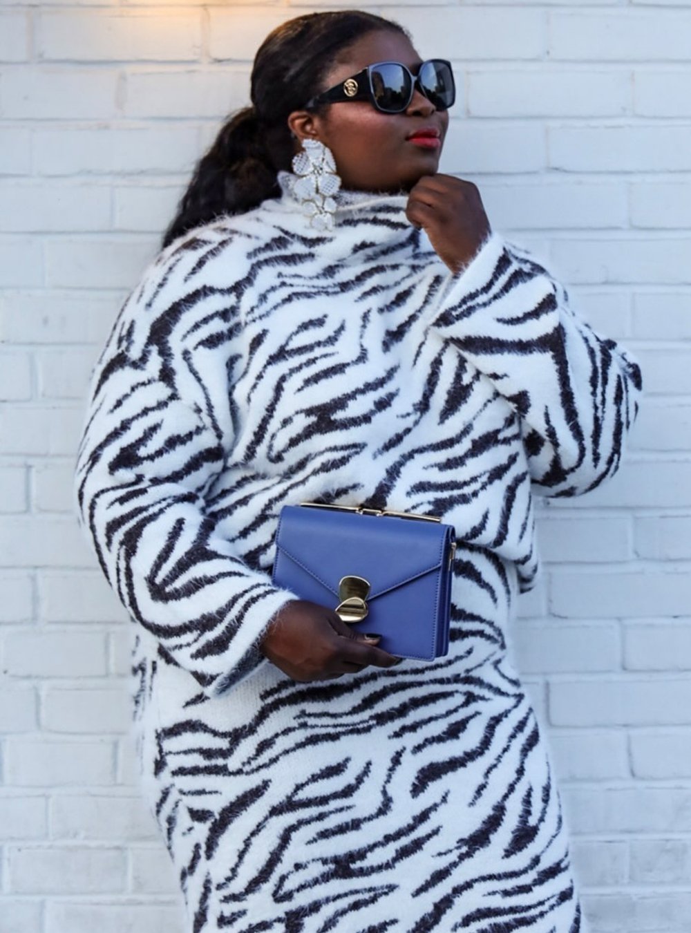 Mulher com suéter e saia de estampa de zebra, segurando uma bolsa azul, usa óculos de sol grandes e brincos chamativos.