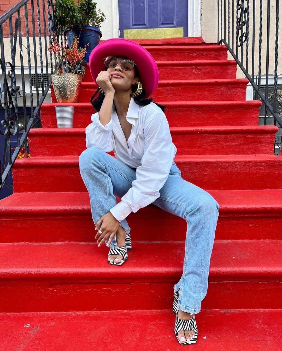 Mulher sentada em escada vermelha, usando chapéu rosa, camisa branca, jeans e sandálias com estampa de zebra.