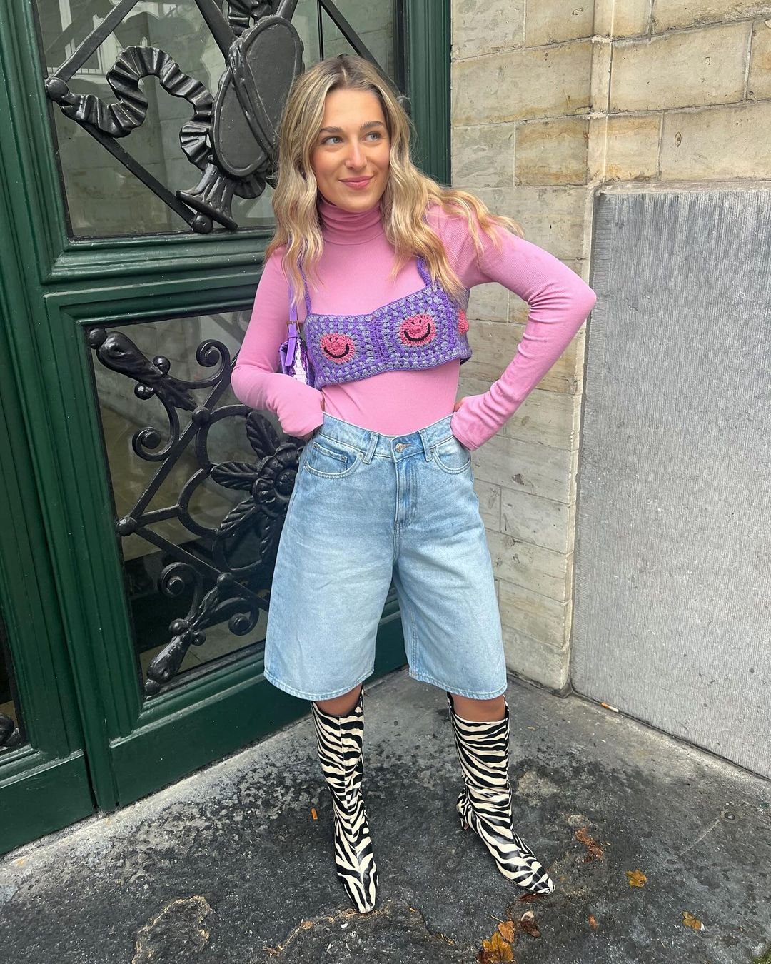 Mulher usando blusa rosa de gola alta, bermuda jeans e botas de cano alto com estampa de zebra, posa em frente a uma porta verde.