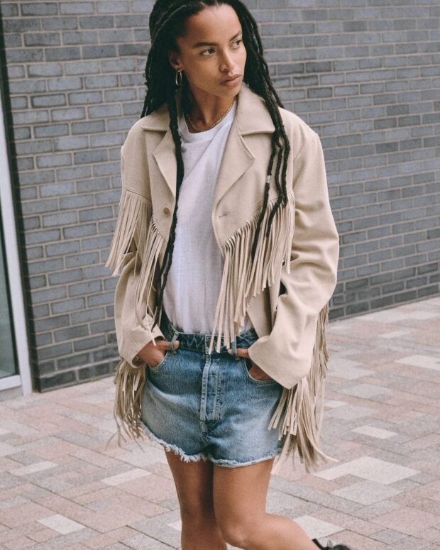 Pessoa usando jaqueta com franjas estilo boho chic, camiseta branca e shorts jeans na rua.