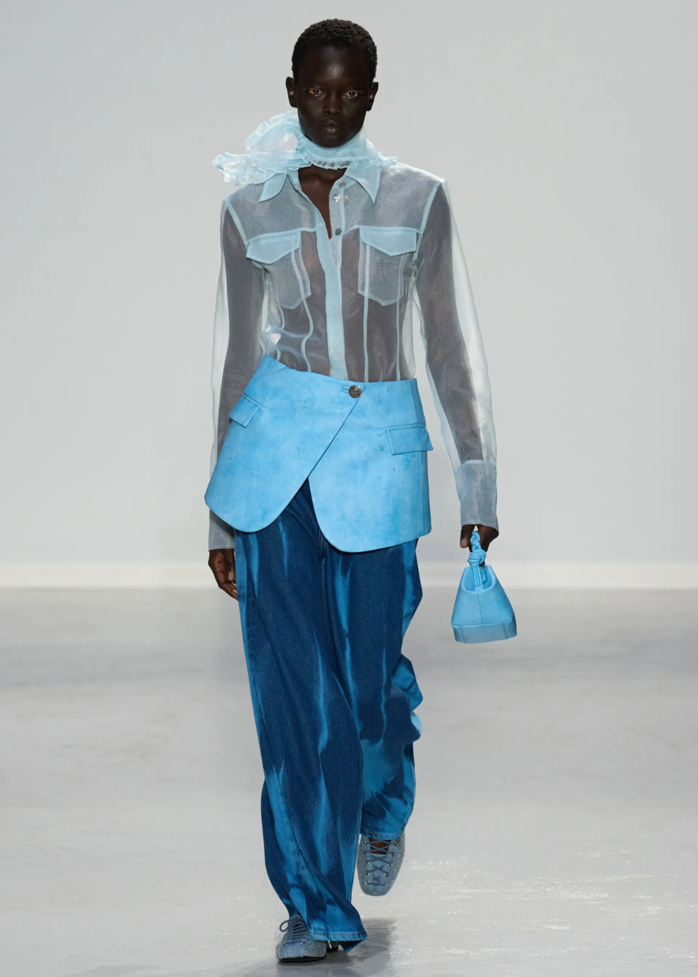 Modelo em desfile de moda com camisa transparente azul, saia estruturada azul claro sobre calça larga azul escuro e tênis combinando. Acessório: pequeno bolso de mão azul e echarpe de organza. Look mistura textura e transparência, destacando tendências modernas.