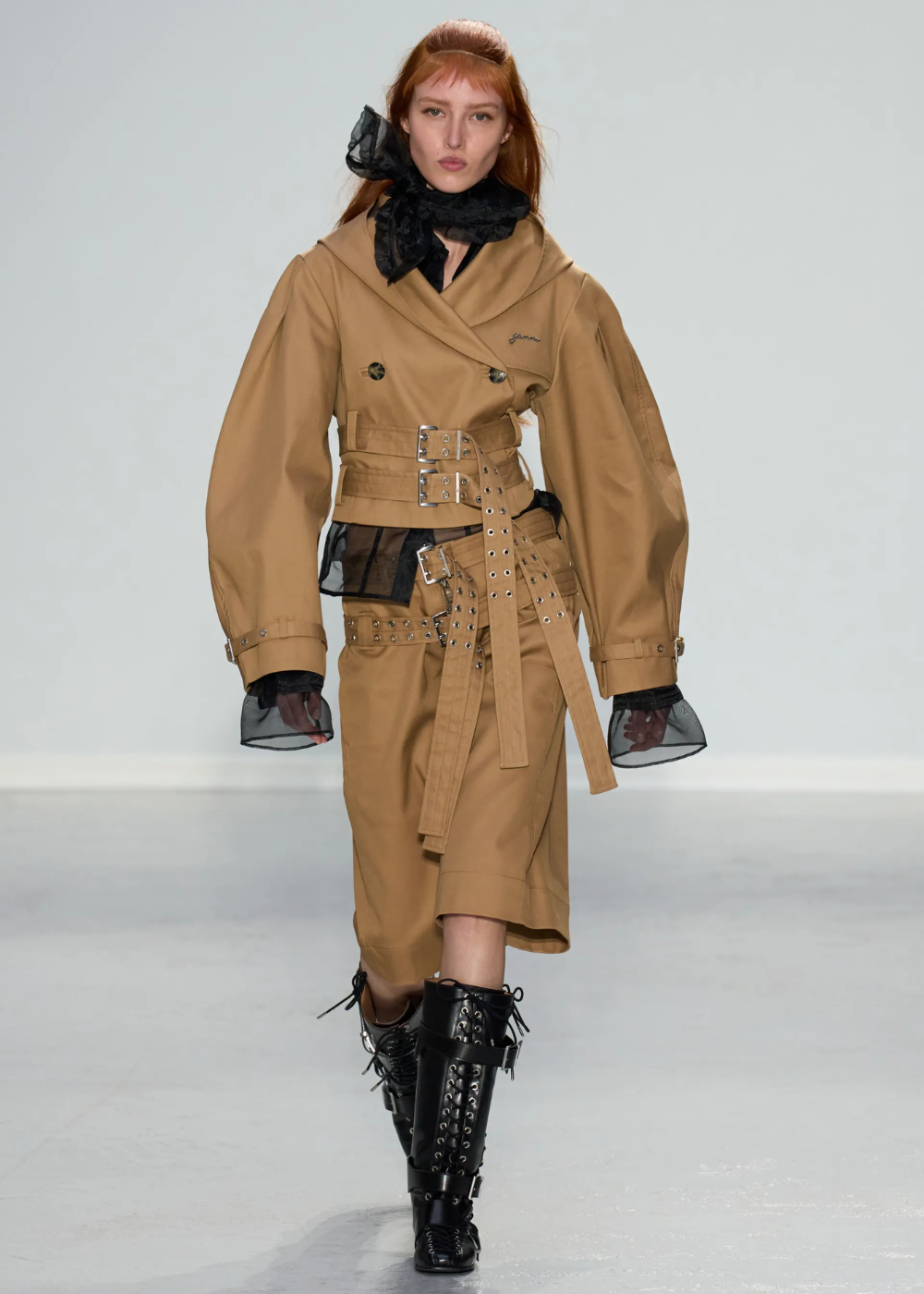 Modelo com trench coat bege, ajustado com cintos e fivelas prateadas. Manga longa com transparências pretas nos punhos e gola. Combina com saia midi e botas pretas de cano alto e amarração, estilo militar. Look ousado e contemporâneo.