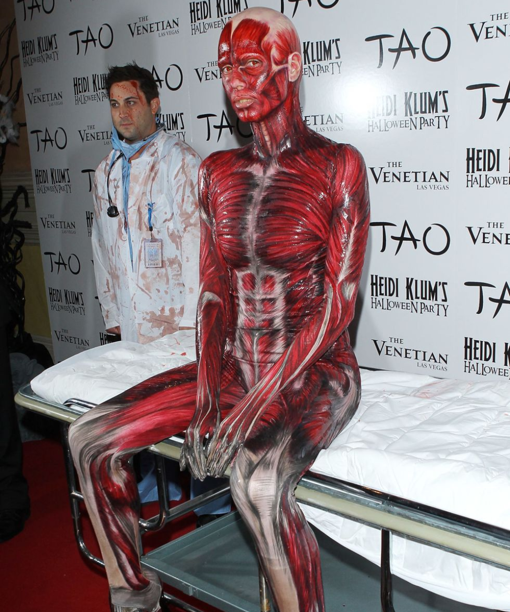 Fantasia temática de Halloween destacando o corpo humano em pintura artística realista, com ênfase em músculos e pele. Ao fundo, traje de laboratório manchado, reforçando o clima de horror e criatividade típica da festa.