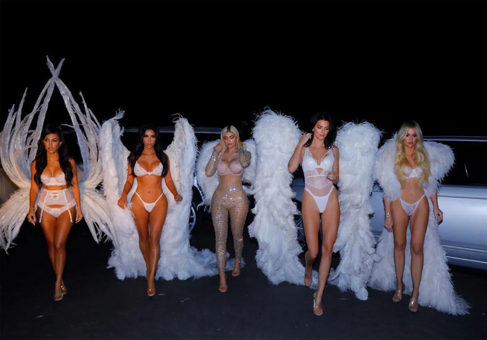 Cinco mulheres vestidas como anjos com lingerie branca e asas enormes. Fantasias das Kardashians.