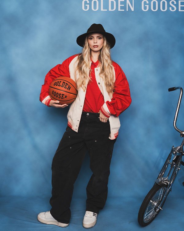 Pessoa vestida com uma jaqueta varsity vermelha e branca, camiseta vermelha, calça jeans larga preta, tênis branco e chapéu preto. Segura uma bola de basquete laranja. Visual mistura tendências streetwear e esportiva.