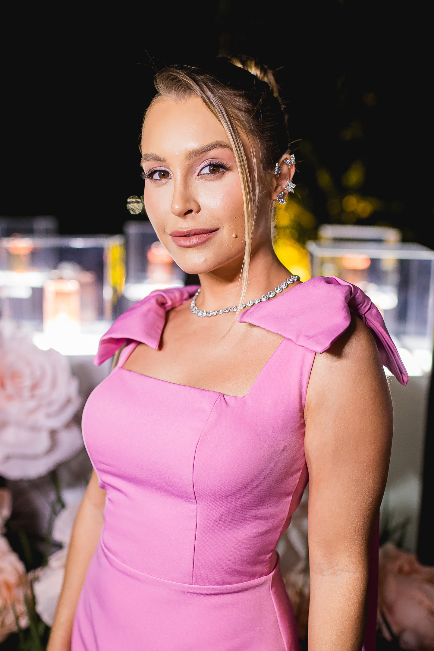 Mulher em vestido rosa brilhante com decoração floral ao fundo, em evento tipo festa do rosa.