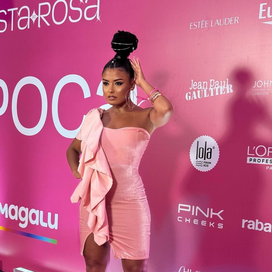 Vestido rosa em mulher posando em evento, tema festa do rosa. Fundo com logotipos de marcas famosas.