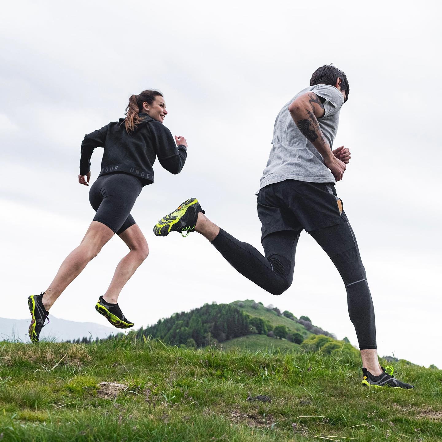 Duas pessoas correndo em ambiente natural. Vestem moda esportiva casual, com leggings pretas, tênis com detalhes em amarelo neon e blusas leves. Estilo foca em conforto e funcionalidade para atividades ao ar livre.