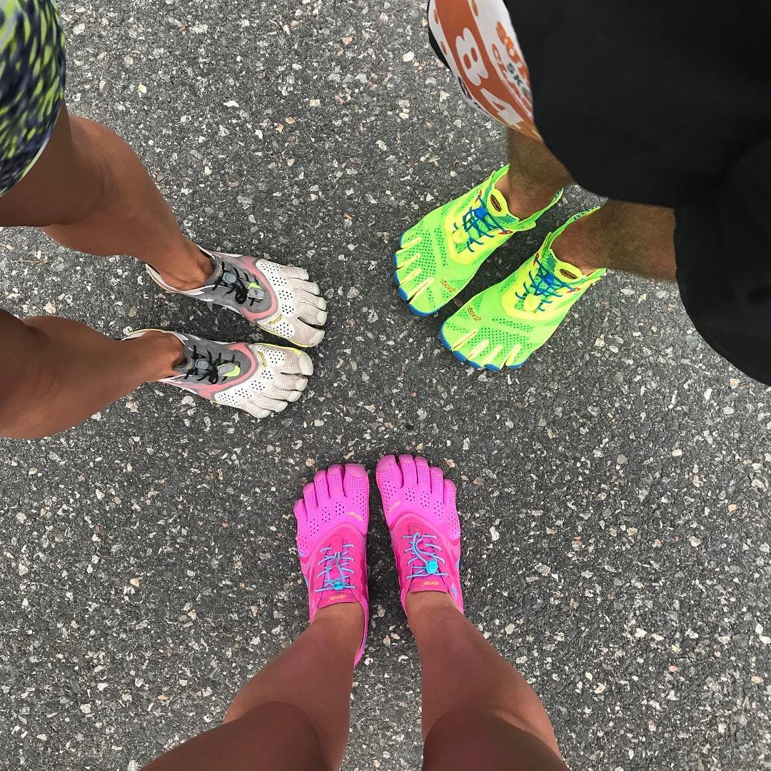 Três pessoas usando sapatos minimalistas com dedos separados, em cores vibrantes: cinza, verde neon e rosa. Tendência de moda esportiva que enfatiza o conforto e a sensação natural ao correr.