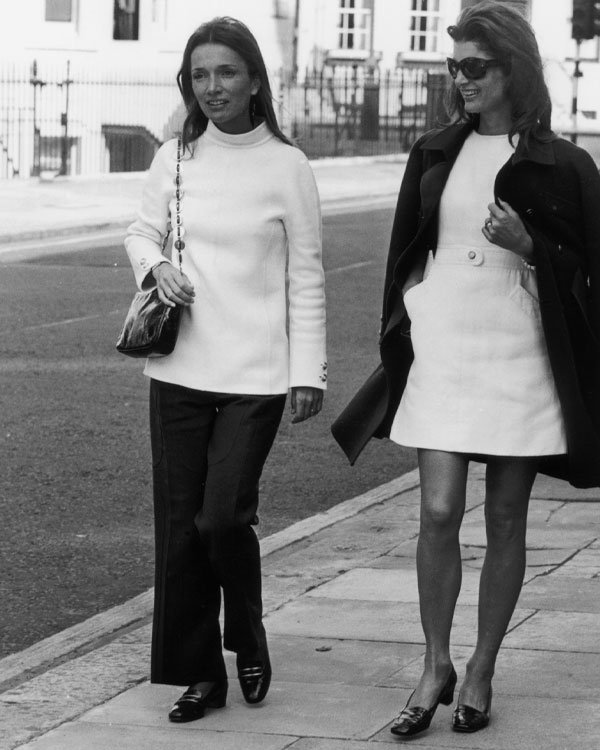 Duas mulheres caminhando em roupa elegante, tendência clássica dos anos 70, na calçada da cidade.