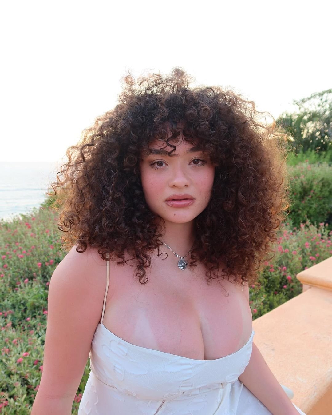 franja no cabelo cacheado em mulher usando vestido branco em área externa com flores.