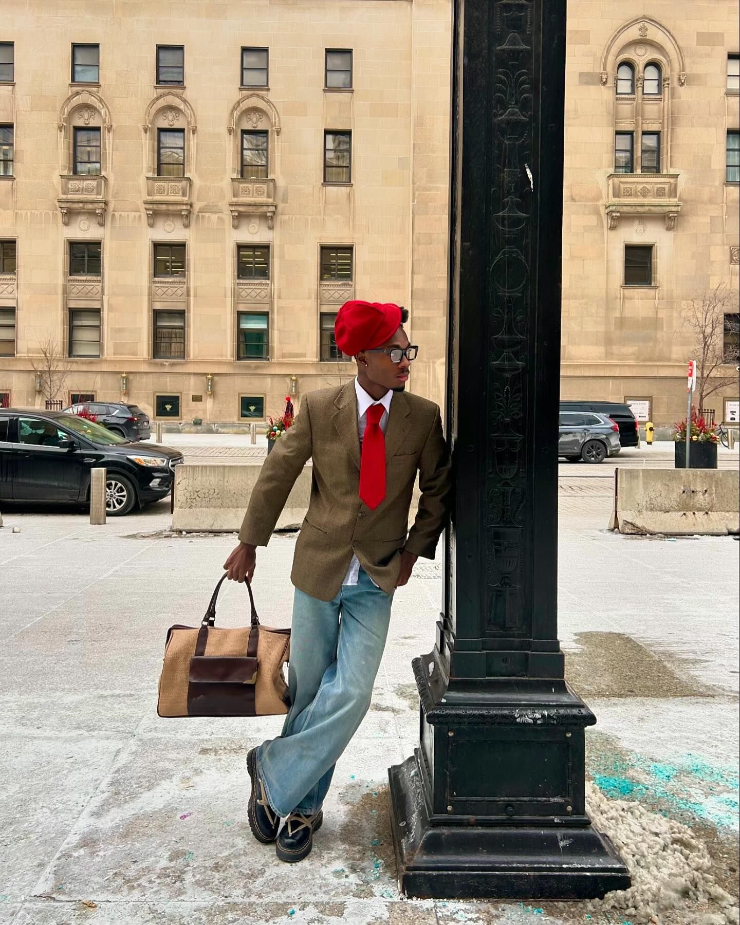 Homem vestido com blazer marrom, calça jeans larga e sapatos de couro estilo oxford. Usa camisa branca, gravata vermelha e boina vermelha, enquanto segura uma bolsa de viagem marrom e bege. O look combina elementos clássicos e modernos.