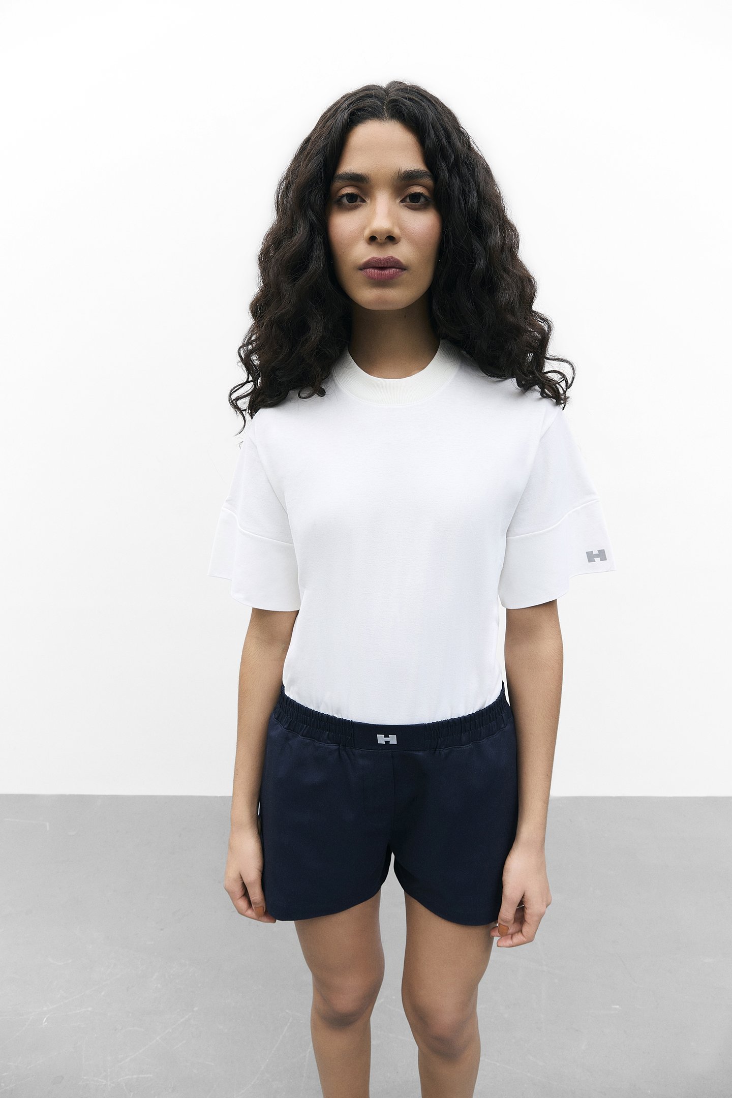 Mulher em pé, usando camiseta branca e shorts pretos, com cabelo cacheado. Fundo branco minimalista.