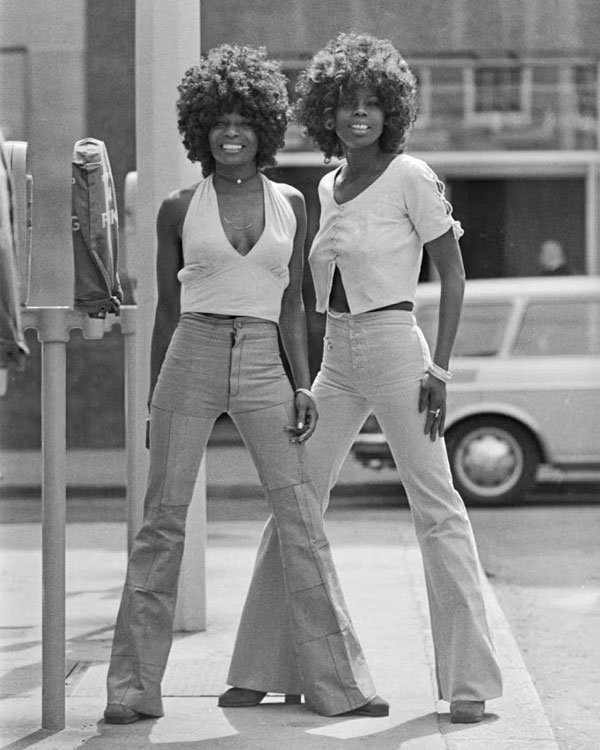 Duas mulheres com visuais dos anos 70 usam calças boca de sino e exibem penteados afro. Elas estão sorrindo na rua.