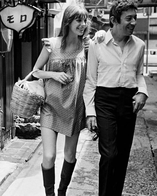 Casal caminhando em rua, vestindo moda dos anos 70. Ela usa vestido curto e botas, ele camisa social e calças.