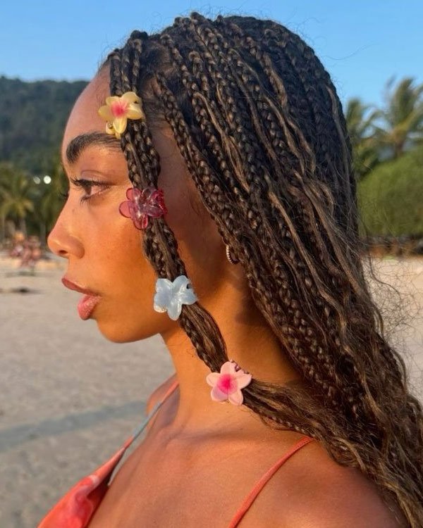 Cabelo trançado adornado com presilhas de flores coloridas, tendência de moda boho e praiana. Roupa de alça fina em tom coral, estilo casual e descontraído, ideal para praia. Fundo com cenário tropical.