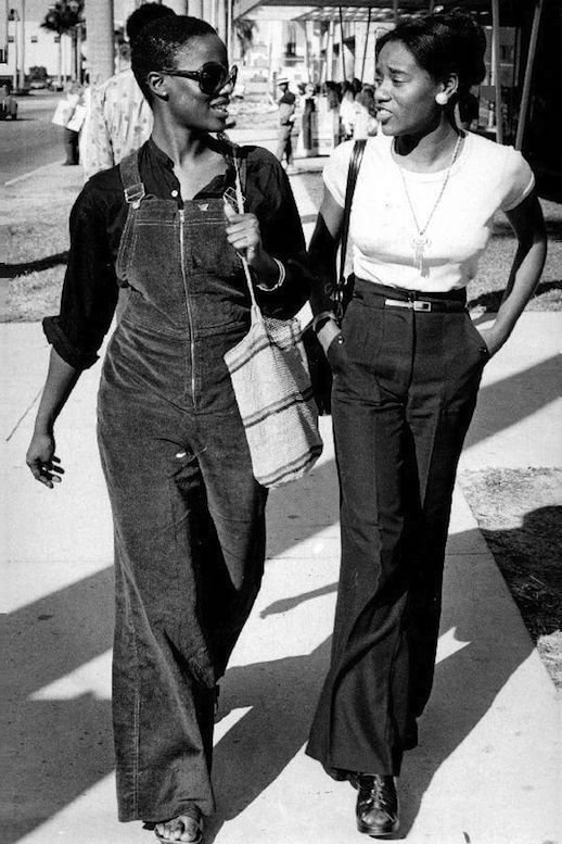 Duas mulheres caminham na rua com roupas de estilo retrô dos anos 70, usando óculos de sol e calças boca de sino.