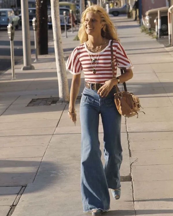 Pessoa caminhando na rua com calça boca de sino e blusa listrada, em um estilo típico dos anos 70.