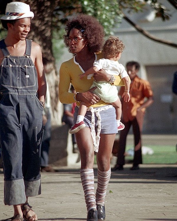 Pessoas passeando com criança, usando moda típica dos anos 70: jardineira, camisa amarela, shorts e meias listradas.